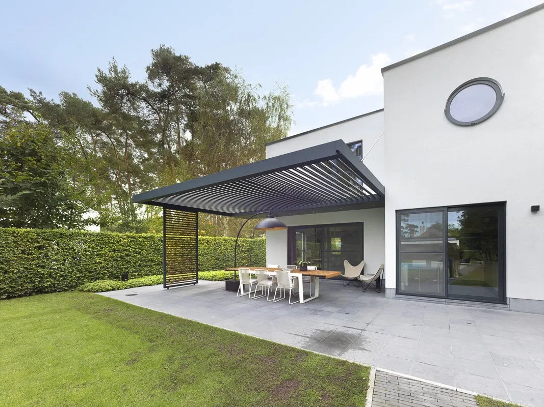 Een modern terras met een zwart pergola dak naast een wit huis met grote ramen en een rond venster. Een houten eettafel en witte stoelen zijn opgesteld onder de pergola, met uitzicht op een netjes onderhouden gazon en een hoge privacyhaag. Hoge dennen op de achtergrond completeren deze rustige buitenruimte.