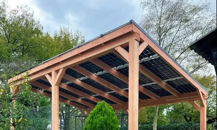 Houten carport met zonnepanelendak in een tuin met groene bomen en een struik.